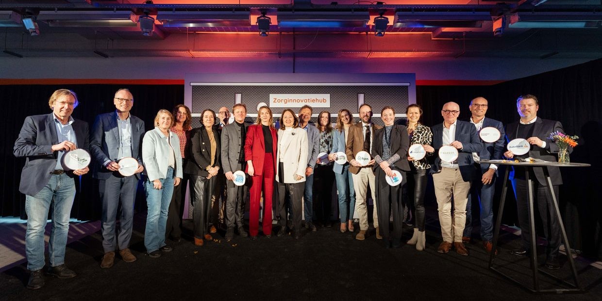 18 mensen, mannen en vrouwen, op het podium die lachend in de camera kijken voor een groepsfoto tijdens de lancering van de zorginnovatiehub