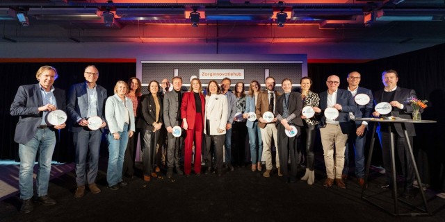 18 mensen, mannen en vrouwen, op het podium die lachend in de camera kijken voor een groepsfoto tijdens de lancering van de zorginnovatiehub